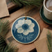 a blue and white ceramic ornament with a white flower design, hanging on a wooden surface. There is also a cup of coffee and some pine branches in the background.