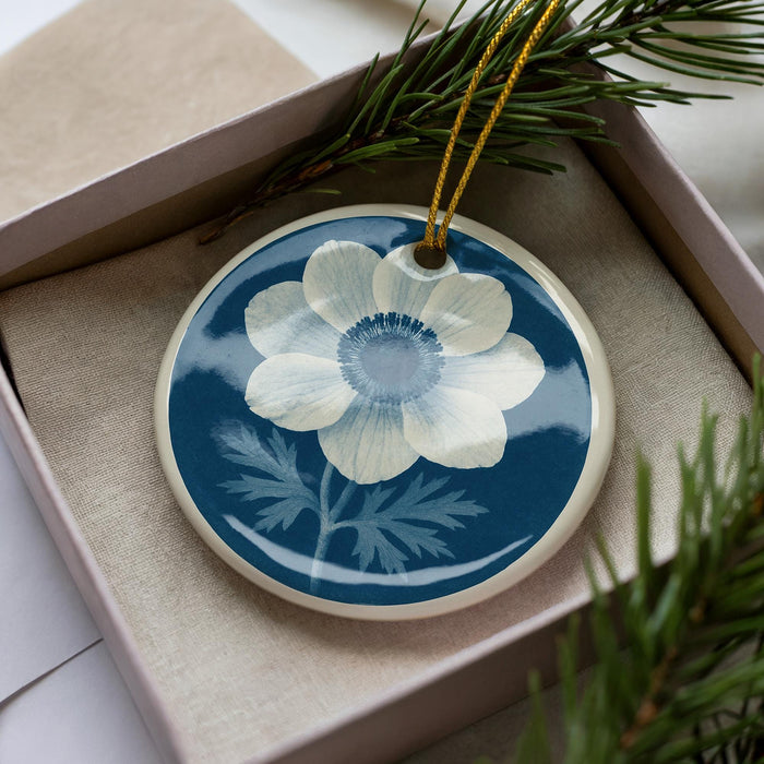 A blue and white ceramic ornament with a white flower design is displayed in a box, surrounded by pine branches.