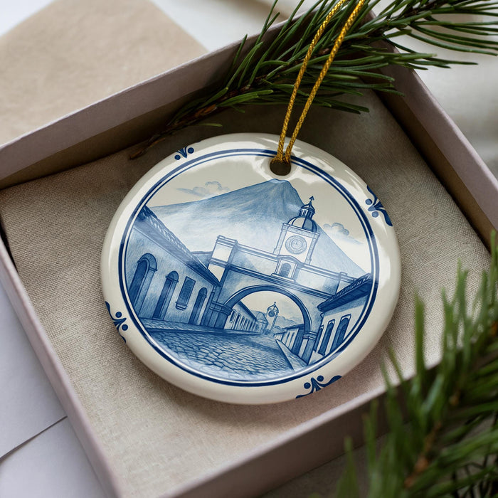A decorative ceramic ornament in the shape of a clock tower, featuring a blue and white design, is displayed in a box with pine branches.