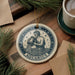 A ceramic ornament featuring the Aquarius zodiac sign, with a figure of a man pouring water, is displayed on a wooden surface alongside a cup of coffee and some pine branches.