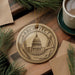 a circular ornament with the word "ARCHITECT" on it, surrounded by architectural tools such as a ruler and a pencil. The ornament is placed on a wooden surface, with pine branches and a cup of coffee nearby.