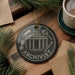 A black ornament with the word "ARCHIVIST" written on it, featuring an illustration of a building and various other objects, is displayed on a wooden surface alongside a cup of coffee and some pine branches.