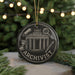 A black ornament with the word "ARCHIVIST" written on it, featuring an illustration of a building and various objects, such as a magnifying glass and a scroll, against a blurred background of greenery and lights.