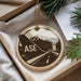 A circular ornament with a graphic design featuring a mountain range and an airplane, along with the text "ASE" on it, is displayed in a box with pine branches.