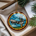 A stained glass ornament in the shape of a plane is displayed in a box, surrounded by pine branches.