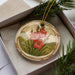A decorative ornament with a floral design, featuring red flowers and green foliage, is displayed in a box with pine branches.