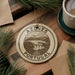 A circular ornament with the text "AZÓRES PORTUGAL" is displayed on a wooden surface, surrounded by pine branches and a cup of coffee.