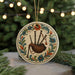 A decorative ornament featuring a piper blowing bagpipe, surrounded by colorful floral designs, is displayed on a table with a blurred background of a Christmas tree.