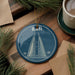 A blue ornament with a drawing of a building, likely a skyscraper, is displayed on a wooden surface alongside a cup of coffee and some pine branches.