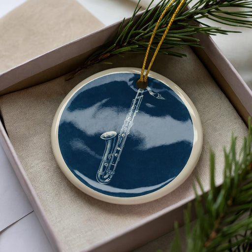 A blue ceramic ornament with a white border, featuring a saxophone design, is displayed in a white box with pine branches.
