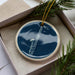 A blue ceramic ornament with a white border, featuring a saxophone design, is displayed in a white box with pine branches.