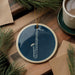 A blue ceramic ornament with a white saxophone design is displayed on a wooden surface, accompanied by a white mug and pine branches.