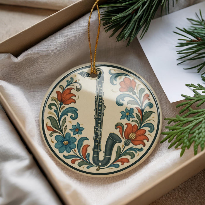 A decorative ornament in the shape of a flute, featuring a floral design, is displayed in a box alongside pine branches and a white card.