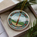 A decorative ornament in a box, featuring a colorful design with a musical instrument and a landscape scene.