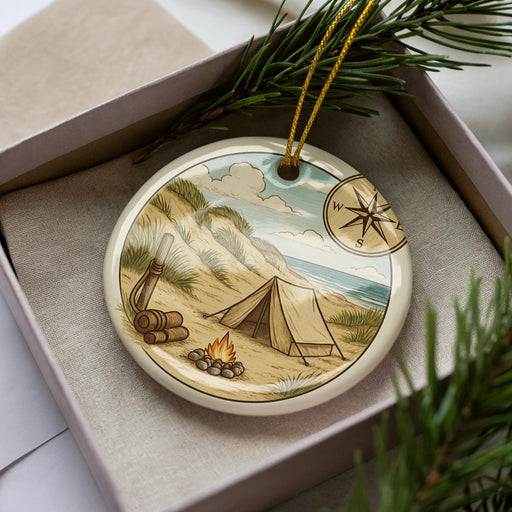 A decorative ornament in a box, featuring a camping scene with a tent, campfire, and compass.