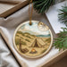 A round ornament with a camping scene, including a tent, campfire, and compass, is displayed in a box with pine branches.