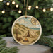 A round ornament depicting a camping scene with a tent, campfire, and a compass, surrounded by pine branches and a blurred background of greenery.