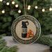 A round ornament with a black spray bottle and a whistle on a tiled border, placed on a surface with pine branches in the background.