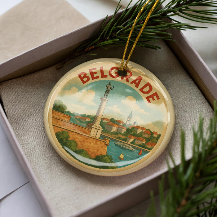 A circular ornament with a picture of a monument and a cityscape, featuring the word "Belgrade" on it, is displayed in a box with pine branches.