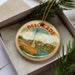 A circular ornament with a picture of a monument and a cityscape, featuring the word "Belgrade" on it, is displayed in a box with pine branches.