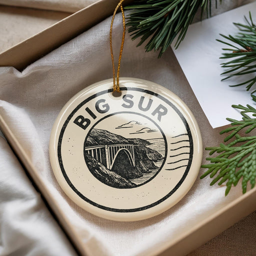 A decorative ornament with the text "BIG SUR" and an illustration of a bridge is displayed in a box with pine branches.