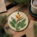 a round ceramic ornament with a tropical flower design, placed on a wooden surface alongside a cup of coffee and some pine branches.