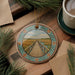 A decorative ornament featuring a wooden bridge over a river, surrounded by pine branches and a cup of coffee on a wooden surface.