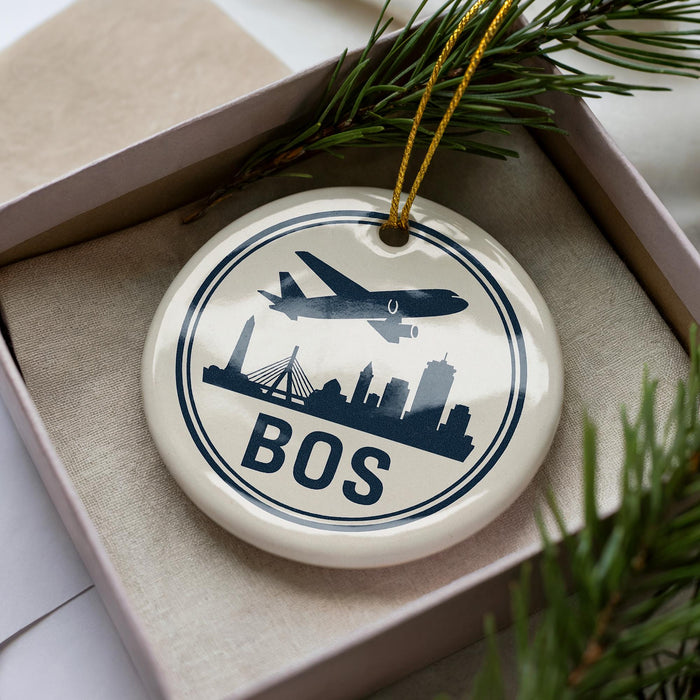 A ceramic ornament with a circular design featuring a silhouette of a city skyline and an airplane, along with the word "BOSTON" written on it. The ornament is placed inside a box, surrounded by pine branches.