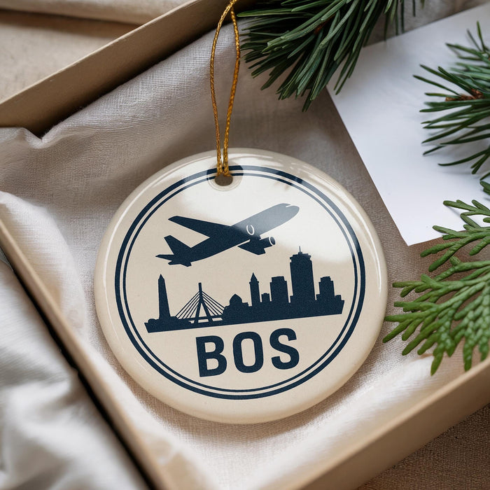 A circular ornament with a silhouette of a plane flying over a city skyline, along with the word "BOS" written on it, is displayed in a box with pine branches.