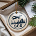 A circular ornament with a silhouette of a plane flying over a city skyline, along with the word "BOS" written on it, is displayed in a box with pine branches.
