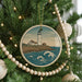 A round ornament on a Christmas tree depicting a lighthouse on a cliff overlooking the ocean.
