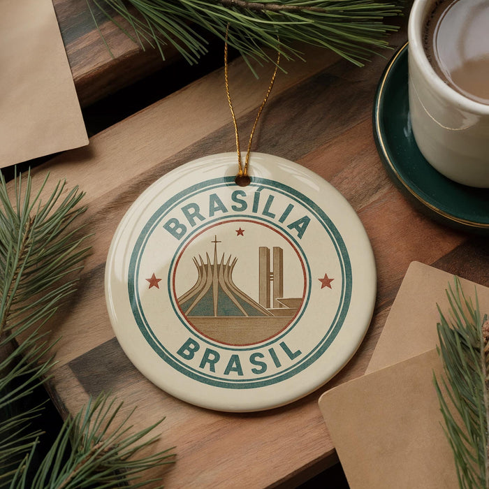 A circular ornament with the text "BRASILIA BRASIL" and an illustration of a church, placed on a wooden surface next to a cup of coffee and some pine branches.