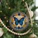 A decorative ornament with a blue butterfly is hanging on a Christmas tree, surrounded by ornaments and lights.