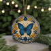 A decorative ceramic ornament featuring a blue butterfly is displayed on a table, surrounded by pine branches and twinkling lights.
