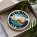 A decorative ornament with a circular design featuring a scenic image of Cabo San Lucas, Mexico, surrounded by pine branches.