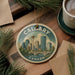A decorative ornament featuring a cityscape of Calgary, Canada, with a bridge and trees, is displayed on a wooden surface alongside a cup of coffee and some papers.