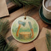 A ceramic ornament featuring a tree stump and two tools, likely a saw and an axe, is displayed on a wooden surface alongside a cup of coffee and some pine branches.