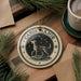 A decorative ornament featuring a dog standing on a rock, surrounded by stars, is displayed on a wooden surface alongside a cup of coffee and some pine branches.
