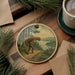 A round ornament featuring a person carrying a large canoe on a path through a forest, with a lake and mountains in the background.