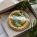 A decorative ornament in a box, featuring a scenic landscape with a tree, water, and buildings.