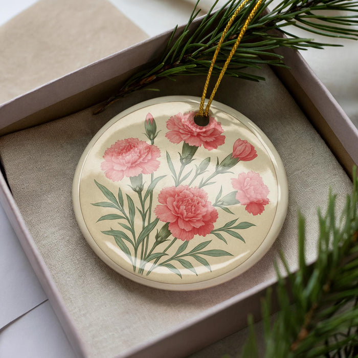 A decorative ceramic ornament with a floral design, featuring pink carnations and green leaves, is displayed in a box with pine branches.