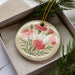 A decorative ceramic ornament with a floral design, featuring pink carnations and green leaves, is displayed in a box with pine branches.