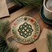 a decorative ornament with a Celtic knot design, surrounded by pine branches and a cup of coffee on a wooden surface.