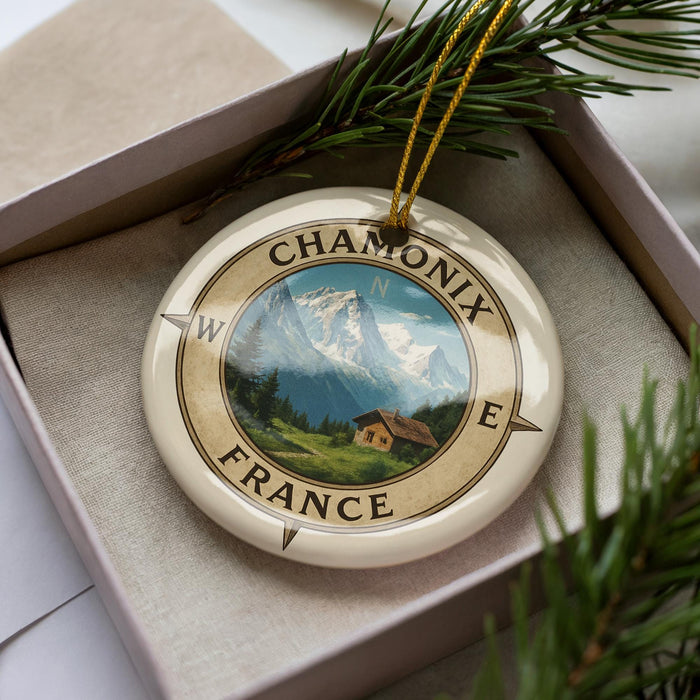 A decorative ornament with a compass design, featuring a mountain landscape and a small cabin, is displayed in a box with pine branches.