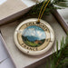 A decorative ornament with a compass design, featuring a mountain landscape and a small cabin, is displayed in a box with pine branches.
