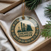 A round ornament with a green and gold design, featuring a chemical engineer's logo, is displayed in a box with pine branches and a white paper.