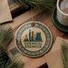 a circular ornament with a design of a laboratory or chemical engineering facility, surrounded by pine branches and a cup of coffee on a wooden surface.