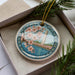 A decorative ceramic ornament featuring a floral design with pink flowers and a blue background, placed inside a white box with a pine branch and greenery.
