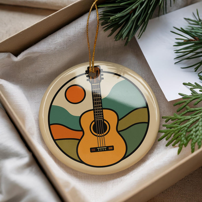 A round ornament with a guitar design is displayed in a box, surrounded by pine branches and greenery.
