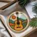 A round ornament with a guitar design is displayed in a box, surrounded by pine branches and greenery.